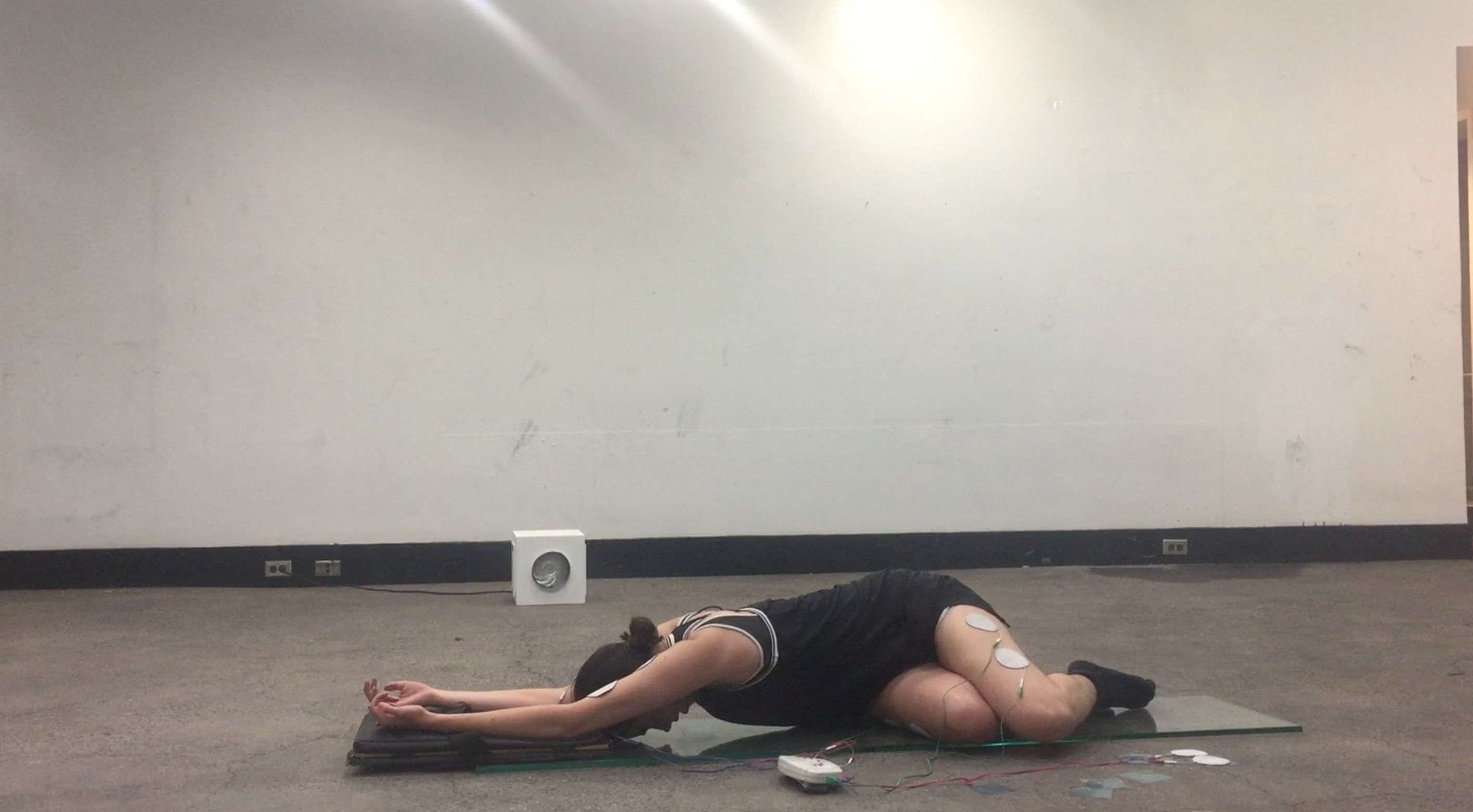 Performers arms stretched out over a leather pad, with their head facing their reflection from the glass below them. Both their legs and arms are patched to the slendertone machine.